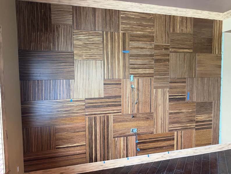 May include: A wall installation composed of rectangular wooden panels featuring a striped pattern of dark brown and light tan wood grain. The panels are arranged in a grid with varying orientations, and small blue tape markers are visible.