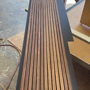 Op de afbeelding: Een rechthoekig houten paneel met verticale, gelijkmatig verdeelde, bruine houten latten. Het paneel ligt op een donkergrijze ondergrond, waarschijnlijk voor montage of presentatie. De houtnerf is zichtbaar.