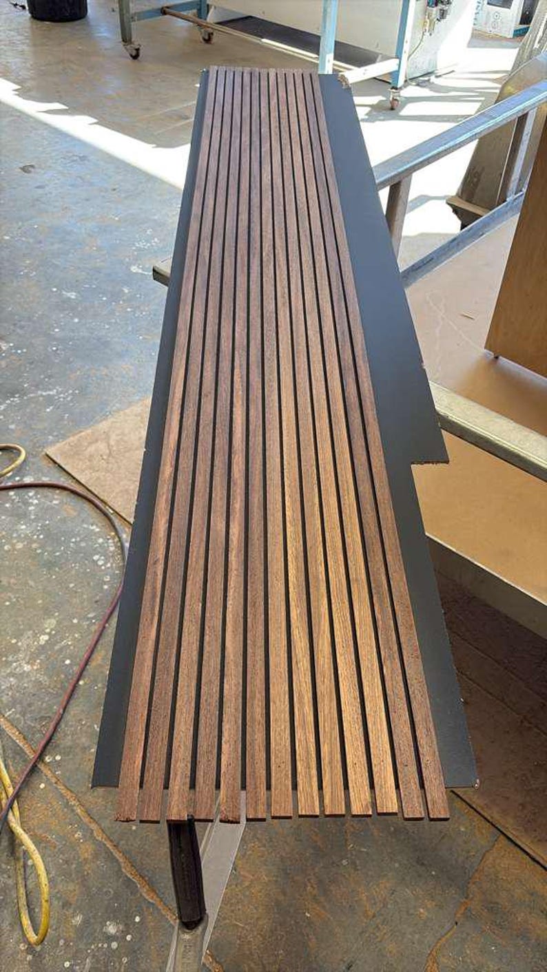 Op de afbeelding: Een rechthoekig houten paneel met verticale, gelijkmatig verdeelde, bruine houten latten. Het paneel ligt op een donkergrijze ondergrond, waarschijnlijk voor montage of presentatie. De houtnerf is zichtbaar.