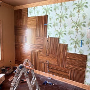 May include: A wall installation featuring rectangular dark brown wood panels with vertical grain patterns arranged in a staggered, geometric layout, partially covering a light blue wallpaper with a green palm leaf and fruit botanical print.