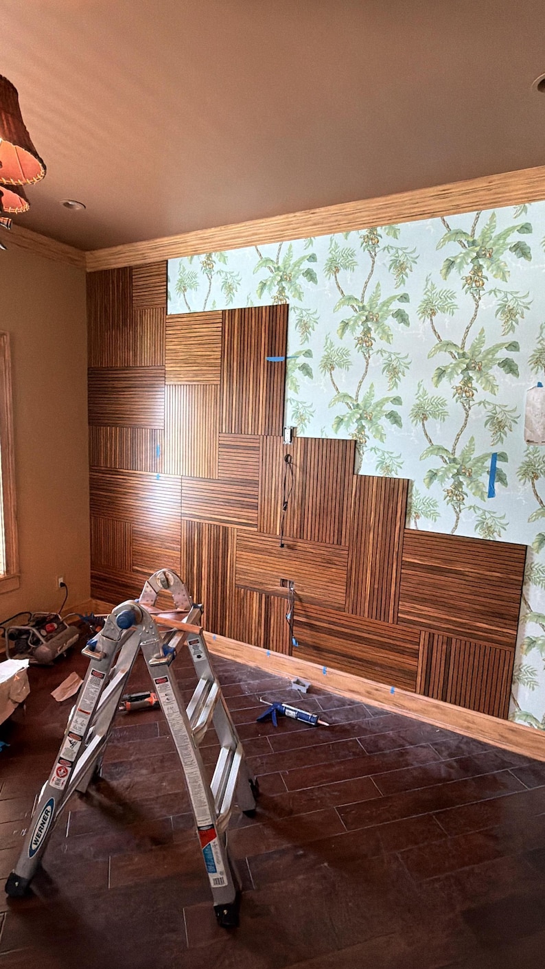 May include: A wall installation featuring rectangular dark brown wood panels with vertical grain patterns arranged in a staggered, geometric layout, partially covering a light blue wallpaper with a green palm leaf and fruit botanical print.