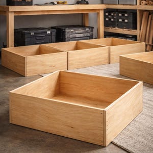 May include: Several unfinished wooden storage boxes of varying sizes are arranged on a workshop floor. The boxes are made of light-colored wood and have a simple, rectangular design. The workshop setting includes tools and storage shelves.