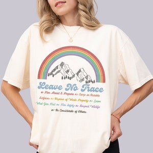 May include: A white t-shirt featuring a rainbow and mountains graphic. The text "Leave No Trace" is printed in blue with a list of principles below in different colours.