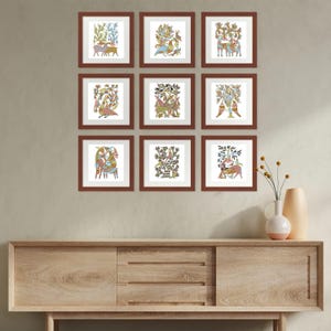 May include: Nine framed art prints with brown frames, each displaying colourful folk art illustrations of animals and plants. The prints are arranged in a grid on a neutral wall above a wooden cabinet. A vase with flowers sits on the cabinet.