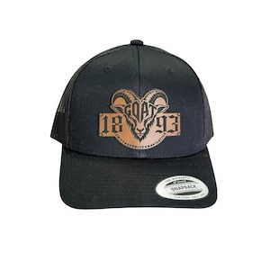 May include: A black snapback cap with a mesh back and a brown leather-like patch on the front. The patch features a goat head graphic, the text GOAT 1893, and the words UNITY SERVICE NAVIGATION. A circular sticker is on the flat brim of the cap.