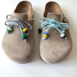 Könnte beinhalten: Ein Paar beigefarbene Wildleder-Clogs mit hellblauen Paracord-Schnürsenkeln, die zu Schleifen gebunden sind. Die Schnürsenkel sind mit bunten Glasperlen in Gelb, Grün, Blau und Weiß verziert. Jeder Schuh hat eine braune Metallschnalle an der Seite.