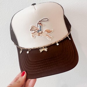 May include: A brown and white trucker hat with a cowboy and horse embroidered patch. The hat features a gold chain with charms, including a bow, a pearl, and a crystal. The brim is brown, and the top is white.