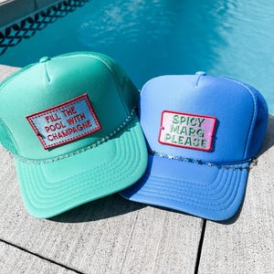 May include: Two trucker hats, one aqua and one blue, with mesh backs and curved brims. The aqua hat has the text "FILL THE POOL WITH CHAMPAGNE" on a patch. The blue hat has the text "SPICY MARG PLEASE" on a patch. Both have a rhinestone trim.