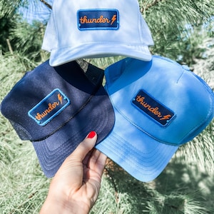 May include: Three trucker hats in white, navy blue, and light blue. Each hat features a rectangular patch with the word "thunder" and a lightning bolt graphic. The hats have a mesh back and a curved brim.