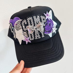 May include: Black trucker hat with the text "COME ON" in a checkered pattern, embellished with a sequined bow, purple smiley face, stars, and a lightning bolt. The hat has a mesh back and a curved brim.
