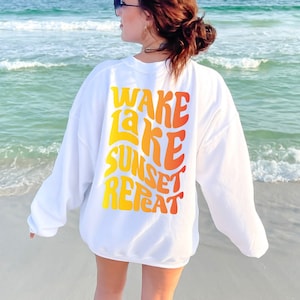 Könnte beinhalten: Weißer Pullover mit einem grafischen Aufdruck auf der Rückseite, der "Wake Lake Sunset Repeat" in einer Retro-Schriftart mit einem Farbverlauf von Gelb, Orange und Rot zeigt.