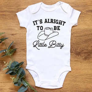 May include: White baby onesie with black text that reads "It's alright to be little bitty" with a cowboy hat graphic.