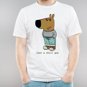 Op de afbeelding: Wit T-shirt met een cartoonillustratie van een bruine hond die een blauwe jeans en een grijze trui draagt. De tekst "Just a chill guy" is onder de illustratie gedrukt.
