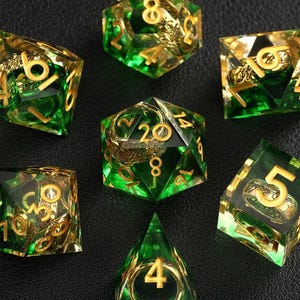 May include: Set of seven polyhedral dice with a green and gold color scheme. The dice are transparent with gold-colored numbers and designs inside. The dice are arranged on a dark surface.