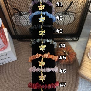 May include: Seven different styles of bracelets with a gold maple leaf charm. Each bracelet is made with different colored stones, including amethyst, blue, black, grey, orange, pink, and red.