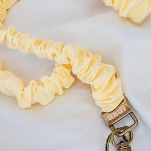 May include: A light yellow fabric lanyard with a textured, gathered design. The lanyard has a bronze-colored metal clasp and key ring. The fabric is a soft, pastel yellow color.