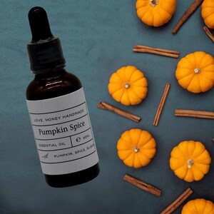 May include: A dark amber glass bottle of Pumpkin Spice essential oil with a dropper, labeled "Love, Honey Handmade." The bottle is surrounded by small orange pumpkins and cinnamon sticks on a teal background. The label reads "Pumpkin Spice Essential Oil e 30ml."