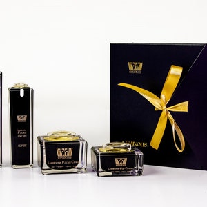 May include: A black gift box with a gold ribbon and the word "LUMINOUS" printed on the front. The box contains a set of four black and gold skincare products: a facial cleanser, a facial serum, a facial cream, and an eye cream.