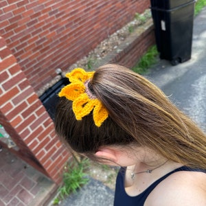 May include: A yellow crocheted sunflower hair tie. The hair tie is worn in a woman's hair, pulled back into a ponytail.