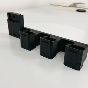 Puede incluir: Un organizador negro impreso en 3D con múltiples compartimentos. El organizador tiene una base rectangular con tres compartimentos cuadrados y una sección más grande a la izquierda. Los compartimentos tienen una letra "T".