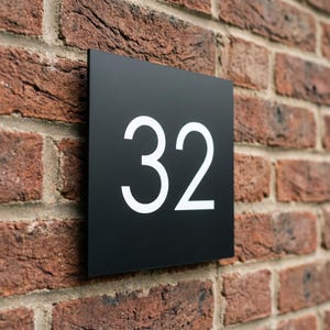 Op de afbeelding: Zwart vierkant huisnummerbord met het witte nummer 32. Het bord is bevestigd aan een rode bakstenen muur. De cijfers zijn groot en duidelijk leesbaar.