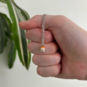 May include: A silver necklace featuring a small pendant. The pendant is a miniature figure in white, orange, and red, with a smiling face. The necklace is held in a hand against a white backdrop.