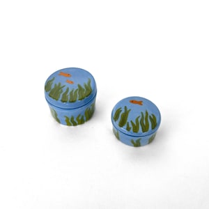 May include: Two round, light blue ceramic trinket boxes with lids. The exterior of both containers features hand-painted green seaweed patterns and small orange fish swimming across the surface, extending from the lids down to the base.