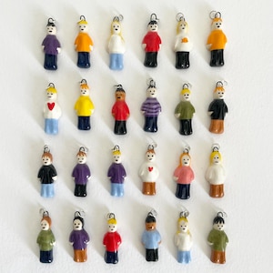 May include: A selection of small ceramic figurines, each representing a person in different outfits. The figurines are in various colours, such as red, yellow, purple, and blue, with black hair and a silver loop for hanging.
