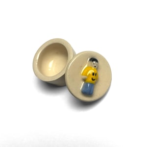 May include: A small, round, off-white ceramic trinket box with a matching lid. The lid features a three-dimensional, hand-painted figurine of a person wearing a yellow shirt and blue pants, with black hair and a simple facial expression.