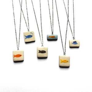 Peut inclure: Plusieurs colliers en céramique faits à la main avec des pendentifs carrés et rectangulaires. Chaque pendentif présente un motif de poisson peint à la main en orange, jaune, bleu et noir. Les pendentifs sont suspendus à de fines chaînes argentées.