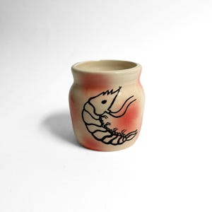 May include: A small ceramic jar with a cream base and a pink-red accent. A black line drawing of a shrimp is on the front. The jar has a rounded shape and a smooth, glossy finish.