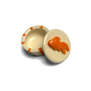 May include: A small, round, off-white ceramic trinket box with a matching lid. The bowl features vertical orange stripes along the exterior rim. The lid is adorned with a raised, three-dimensional orange goldfish figure centered on the top surface.