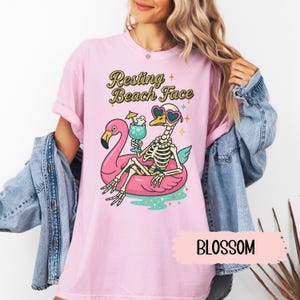May include: A light pink t-shirt with the text "Resting Beach Face" above a skeleton wearing heart-shaped sunglasses and floating on a pink flamingo float. The shirt also has the word "BLOSSOM" on it.