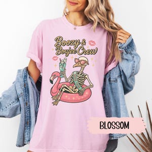 May include: A light pink t-shirt with the text "Bozy & Boujee Crew" and a graphic of a skeleton wearing sunglasses and a "Bride Tribe" sash, floating on a pink flamingo floatie. The shirt also features a cocktail and lipstick graphics.