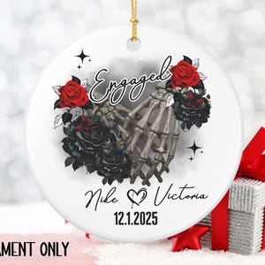 May include: White ceramic ornament with a gold string, featuring a skeleton hand design holding a ring, surrounded by red and black roses. The word "Engaged" is written above the hands, with names and a date below.
