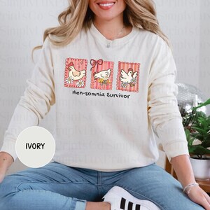 May include: Ivory-colored sweatshirt with the text "Hen-somnia Survivor" and three framed chicken illustrations. Each frame is pink with a scalloped edge and features a different chicken design with floral accents. The sweatshirt is long-sleeved.