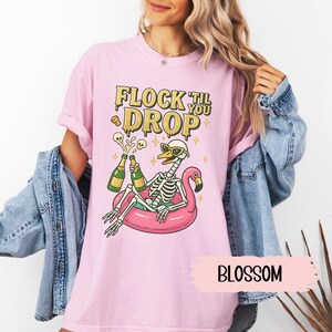 May include: Pink t-shirt with the text "FLOCK TIL YOU DROP" in gold lettering. The graphic features a skeleton wearing sunglasses, relaxing on a pink flamingo floatie, and holding champagne bottles. The word "BLOSSOM" is written on a pink banner.