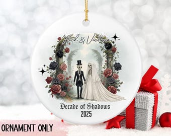 May include: A white ceramic ornament with a gold string, featuring a skeleton couple in wedding attire. The design includes black and red roses, the names "Nick & Victoria," and the text "Decade of Shadows 2025."