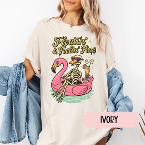 May include: Ivory-colored t-shirt with a graphic of a skeleton relaxing on a pink flamingo floatie, wearing sunglasses and holding a cocktail. The text "Floatin' & Feelin' Fine" is above the graphic. The word "IVORY" is below the graphic.