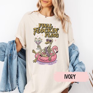 May include: Ivory-colored t-shirt with a graphic featuring a skeleton on a pink flamingo float, holding a cocktail. The text "FINAL FLOCKIN' FLING" is in gold lettering. The shirt is a relaxed fit.