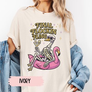 May include: Ivory-colored t-shirt with a graphic of a skeleton flamingo wearing sunglasses and holding a cocktail, floating on a pink pool float. The text "FINAL FLOCKIN FLING" is above the flamingo.