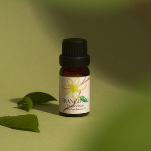 May include: A small brown glass bottle with a black cap, labeled "FRANGIPANI Peppermint 100% Pure Essential Oil". The bottle is sitting on a green surface with green leaves.