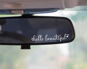 May include: A car's rearview mirror with the words "hello beautiful" in white script. The text is accompanied by small heart shapes. The mirror is set against a blurred background of a car's interior.