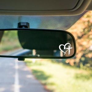 May include: A white heart and cross decal is affixed to a car's rearview mirror. The decal is a simple, minimalist design, with the heart and cross appearing to be hand-drawn. The background is blurred, suggesting the car is in motion.