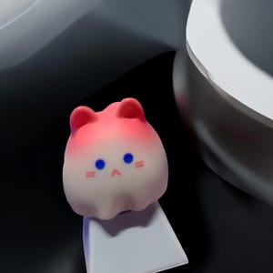 May include: A pink and white gradient cat-shaped keycap with blue eyes and a small pink nose. The keycap is sitting on a white keycap base.