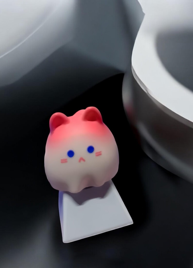 May include: A pink and white gradient cat-shaped keycap with blue eyes and a small pink nose. The keycap is sitting on a white keycap base.
