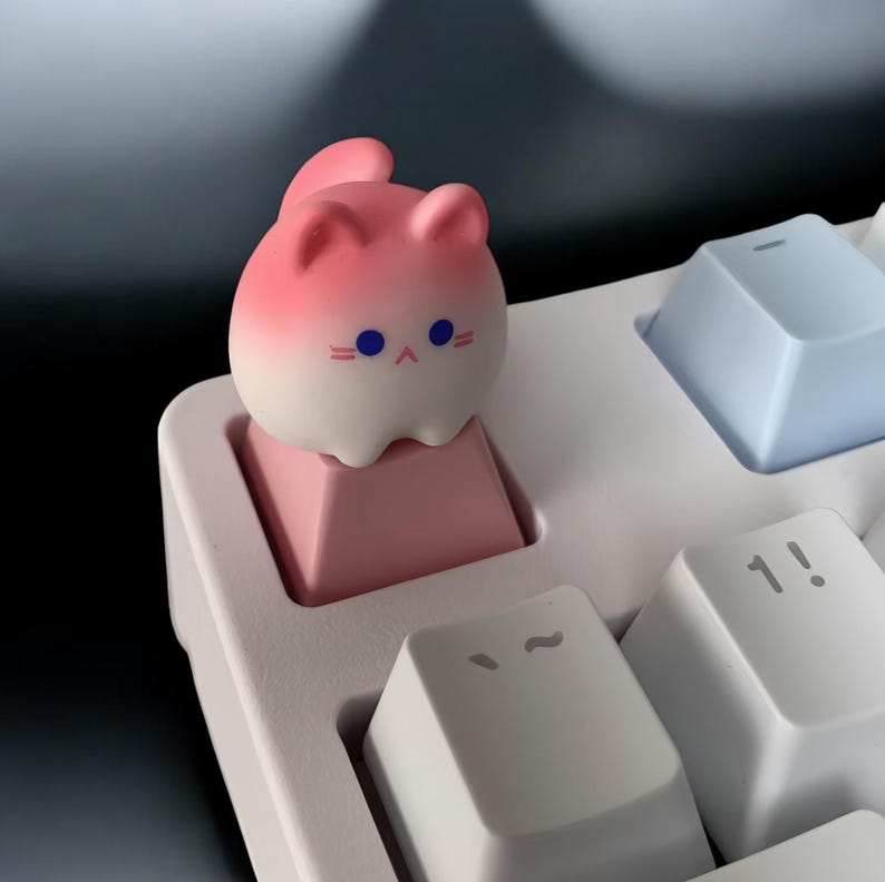 May include: A pink and white cat-shaped keycap with blue eyes sits on a pink keycap on a white keyboard.