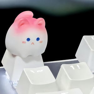 May include: A white and pink cat-shaped keycap with blue eyes and a pink blush. The keycap is sitting on a white keyboard.