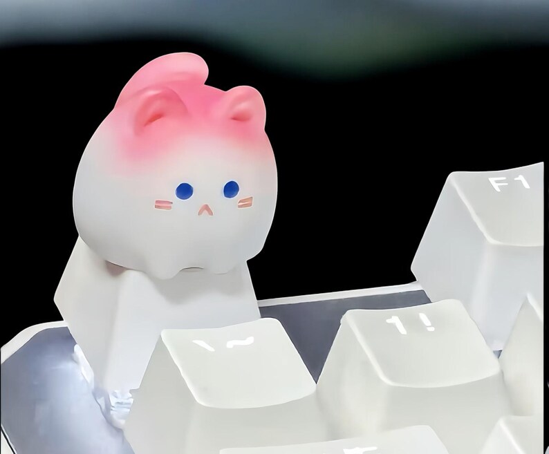 May include: A white and pink cat-shaped keycap with blue eyes and a pink blush. The keycap is sitting on a white keyboard.