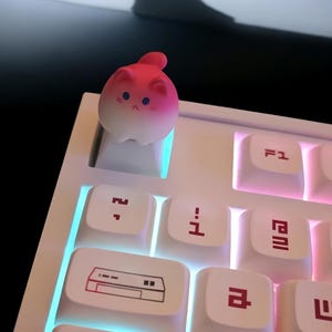 May include: A white keyboard with pink and blue backlighting. A pink and white cat-shaped keycap is on the 'F' key.